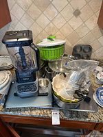 Ninja blender with blender base and clear pitcher, assorted pots, pans, plates, salad spinner, muffin tin, and kitchen gadgets grouped on countertop.