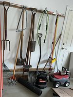 Full view of all garden tools and equipment lined up by the wall, showing rakes, shovels, saw, blower, riding toy, and storage bag.