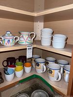 Corner shelf displaying Pier 1 egg coddlers with lids and spoons, various colorful mugs including small and large floral design mugs, and Italian cream and sugar set.