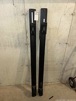 Front view of two vertical black roof racks with locking mechanisms, standing against a concrete wall.