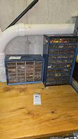Two blue metal storage cabinets filled with assorted screws and fasteners on a wooden bench against a basement wall with visible piping.