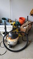 Garage corner view with Ridgid shop vac, antique bench, orange bucket, green watering can, metal scoop, and other garden supplies visible