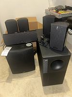 Wide shot showing six Definitive Technology Pro Cinema surround speakers and one Bose subwoofer grouped together on floor.