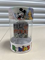 Front view of packaged Rubik's Cube showing Disney characters Mickey Mouse and Simba on the packaging and cube face showing parts of character artwork and icons.