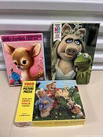 Front view of three puzzle boxes: Little Darlins with a deer, MB Cassette with Miss Piggy and Kermit, TUCO teddy bears puzzle.