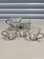 Four clear glass espresso cups arranged with original box in back, showing stainless steel handles and bases.