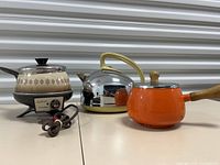 Three vintage kitchen items placed side by side on white surface: Sunbeam fondue pot on left with control knob, shiny chrome General Electric tea kettle in center with yellow handle, and bright orange enamel pot with wooden handle and lid on right.