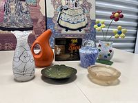 Photo showing entire lot on white table including multiple vases, decorative dishes, floral vase with metal flower ornament, and boxed vintage candle holder.