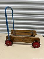 Side view of the wooden trolley with a blue metal handle and red metal wheels on white background.