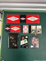 Top-down view of all game items laid out on a green surface including two ACME Deluxe Cribbage Boards, a set of playing cards, bridge card set, trivia cards, and two score pads.