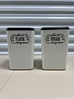 Front view of both vintage canisters labeled Flour and Sugar, showing their rectangular form and black tops.
