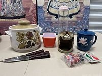 Photo of all items on display including casserole dish, knives, Pyrex container, pottery face piece, enamel mug, straw dispenser, and spools.