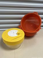 Three vintage plastic kitchen items: two orange strainers and one yellow and white salad spinner