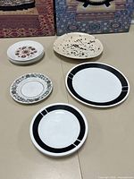 Top view of lot showing all plates arranged on table: floral small Corelle plates stacked, single black filigree Corelle plate, large black and white Corelle plate, and Manitou Brindley England plate with speckled black and cream pattern.