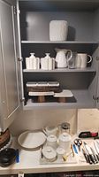 Cabinet shelves displaying ribbed vase on top, cluster vases, white pitchers, clear glass vase, beige pitcher on middle shelf, woven basket with bowls and cake stands on lower shelf and countertop items