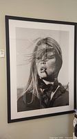 Framed black and white photograph art piece of a woman smoking a cigar with windblown hair, hung on a wall.