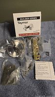 Photo of one Taymor Builder Series keyless one-sided deadbolt lock with all components spread out, including deadbolt mechanism, strike plate, mounting screws, and instructions on a blue cloth background, along with the product box visible.