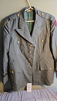 Full front view of military green jacket on hanger showing buttons, pockets, shoulder epaulets, and one sleeve patch.