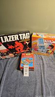Three boxed games: Lazer Tag, KerPlunk, and Monopoly Travel Edition arranged side by side on a fabric surface.