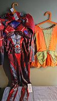 Two youth costumes hanging on orange hangers against a gray wall. Left is a red superhero costume with muscle and armor details. Right is an orange and gold vintage style dress with decorative trim.