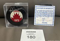 Photo shows the Paul Henderson autographed hockey puck in its clear acrylic holder next to the certificate of authenticity.