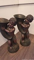 Pair of composite statues depicting cherubic Cupid figures holding large horn-shaped elements, standing on round bases, shown side by side on a wooden surface.