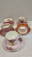 Five vintage style teacup and saucer sets arranged on white surface, showcasing various pink tones, floral patterns, and gold trim details.