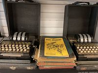 Two Enigma machines side by side with stack of coding books between them