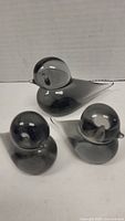 Photo showing three smokey grey glass bird figurines on a white background, viewed from a diagonal angle highlighting their rounded bodies and pointed tails.