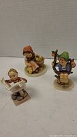 Three porcelain figurines about 3 inches high, two marked Hummel, one unmarked. Included a boy reading newspaper, girl with basket, boy with green hat.