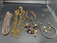 Overview of six necklaces displayed on a black surface including a colorful bead and coin charm necklace, gold-tone chain, twisted silver-tone necklace, and multi-strand metallic mesh necklace.