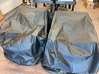 Two large black patio chair covers displayed over patio chairs indoors on wooden flooring.