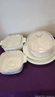 Photo showing white Pyrex casserole bowls with clear glass lids and white Corelle plates, all placed on a purple cloth