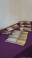 Photo showing 12 knives with ornate decorated gold-tone handles, 11 forks, dessert forks, and cutting utensils arranged on a purple cloth.