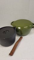 Black cast iron pot and green enamel Copco pot with lids, separate wood handle laid out.