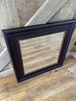 Front view of the square mirror showing dark wooden frame and beveled glass reflecting wood floor.