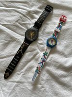 Two Olympic commemorative watches laid out on white fabric, showing both watches fully, one black and one white with colorful athlete figures.