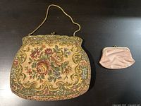 Photo showing an antique floral needlepoint purse with ornate metal frame and handle next to a small pink fabric coin purse with metal frame, both placed on a dark surface.