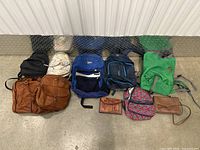 Image showing all 8 bags and backpacks arranged on the floor including green outdoor backpack, dark blue multi-compartment backpack, royal blue backpack, brown purses, white patterned backpack, small black bag, and multicolor patterned backpack.