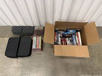 Box containing a collection of Blu-Ray and DVD movies along with two black padded cases and a stack of CDs or DVDs.