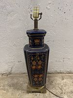 Front view of the tall table lamp showing dark blue ceramic body with floral decoration and brass base, unplugged and unlit, against a grey wall.