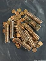Photo showing a pile of loose copper pennies alongside multiple rolls of pennies in transparent coin wrappers on a dark surface.