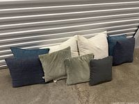 Photo of eight cushions arranged in two rows against a metal wall. Cushions are varied in color including white, blue, green/gray, and dark gray.
