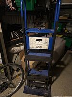 Blue Penske two-in-one hand truck dolly with label visible, stored in a crowded area with other items around it.