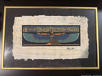 Framed image of Egyptian goddess depicted with blue and green wings seated with arms outstretched, displayed on a papyrus sheet with rough edges, black mat and gold frame.