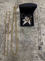 Jewelry lot displaying two gold-tone rope chain necklaces, silver-tone necklace with pendant, and a black box containing rings and a leaf brooch.