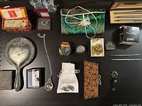 Overview of various vintage jewelry, watch parts in jar, hand mirror, pen set, and accessories arranged on dark surface