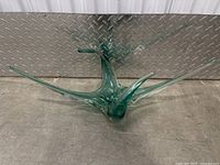 Full view of large green glass sculpture sitting on a floor with a metallic diamond plate background showing the full starburst shape with multiple long spikes.