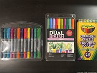 Photo showing a set of 12 dry erase markers, a pack of 10 dual brush pens, and a 24-count box of Crayola colored pencils on a black surface.
