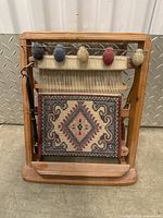 Front view of miniature wooden loom with colored yarn balls and woven geometric tapestry.